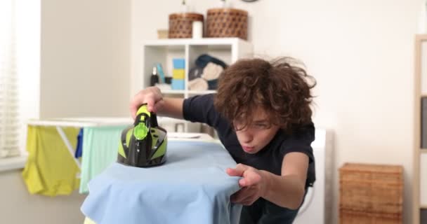 Hard-working teenager wearing blue shirt, irons on ironing board, takes care of clothes, performs household chores in laundry room — Stock Video