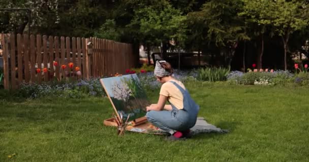 クリエイティブガールは、キャンバスを作成するためのツールを使用して、パレットやブラシを保持し、庭で美しい絵を描いています。アーティストとアートワークのコンセプト — ストック動画