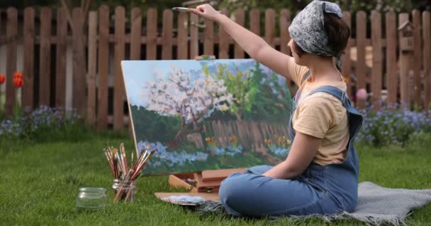 Fata creativă pictează o imagine frumoasă în grădină, ținând o paletă și o perie, folosind instrumente pentru a crea o pânză. Artiști și concept de artă — Videoclip de stoc