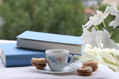 Kahve zamanı konsepti, bir fincan kahve, güzel beyaz orkide, ev yapımı kurabiyeler ve beyaz bez üzerine mavi kitaplar, açık hava çekimi.