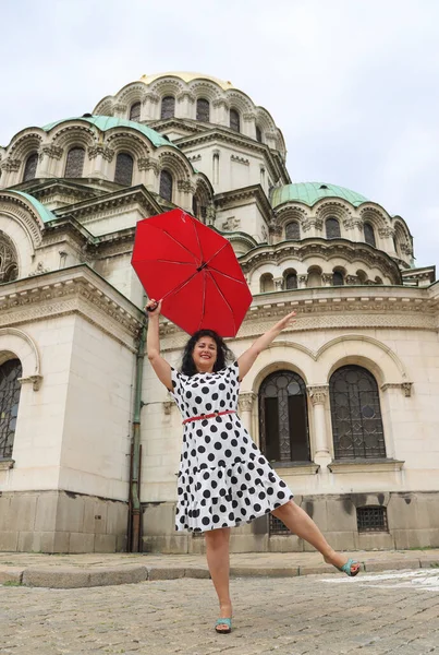 A beautiful brunette woman in a spotted dress holding a red umbrella posing in front of Alexander Nevsky Cathedral in Bulgaria