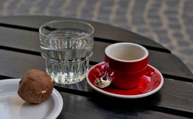 Kırmızı bir fincan üzerinde kahve duran küçük kırmızı bir fincan. Yanında berrak bir bardak su ve beyaz tabakta top şeklinde çikolatalı kek var. Sokakta bir kafe yakınında koyu ahşap bir masa duruyor..