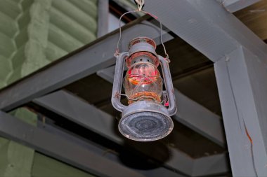 Yanmış bir fener ahşap bir kirişe asılı. Metal ve cam gölgelerden yapılmış arka sokak ışıklandırması tepe örtüsüne asılı duruyor. Akşam erken saatlerde.