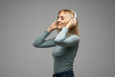 Mavi tişört ve gözlüklü, beyaz kulaklıklı, müzik dinleyen ve stüdyoda gri arka planda gülümseyen güzel sarışın kadın. Rahatlamak ve eğlenmek. Yaşam biçimi.