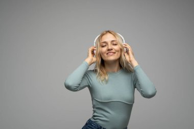 Mavi tişört ve gözlüklü, beyaz kulaklıklı, müzik dinleyen ve stüdyoda gri arka planda gülümseyen güzel sarışın kadın. Rahatlamak ve eğlenmek. Yaşam biçimi.