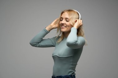 Mavi tişört ve gözlüklü, beyaz kulaklıklı, müzik dinleyen ve stüdyoda gri arka planda gülümseyen güzel sarışın kadın. Rahatlamak ve eğlenmek. Yaşam biçimi.