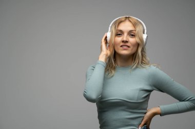 Mavi tişört ve gözlüklü, beyaz kulaklıklı, müzik dinleyen ve stüdyoda gri arka planda gülümseyen güzel sarışın kadın. Rahatlamak ve eğlenmek. Yaşam biçimi.