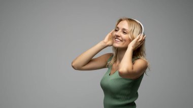 Yeşil tişört ve gözlüklü, beyaz kulaklıklı, müzik dinleyen ve stüdyoda gri arka planda gülümseyen güzel sarışın kadın. Rahatlamak ve eğlenmek. Yaşam biçimi.