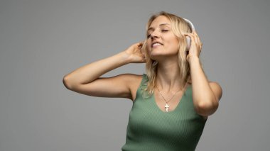 Yeşil tişört ve gözlüklü, beyaz kulaklıklı, müzik dinleyen ve stüdyoda gri arka planda gülümseyen güzel sarışın kadın. Rahatlamak ve eğlenmek. Yaşam biçimi.