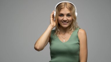 Yeşil tişört ve gözlüklü, beyaz kulaklıklı, müzik dinleyen ve stüdyoda gri arka planda gülümseyen güzel sarışın kadın. Rahatlamak ve eğlenmek. Yaşam biçimi.