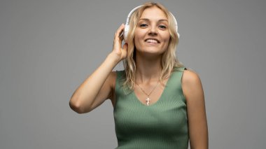 Yeşil tişört ve gözlüklü, beyaz kulaklıklı, müzik dinleyen ve stüdyoda gri arka planda gülümseyen güzel sarışın kadın. Rahatlamak ve eğlenmek. Yaşam biçimi.