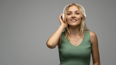 Yeşil tişört ve gözlüklü, beyaz kulaklıklı, müzik dinleyen ve stüdyoda gri arka planda gülümseyen güzel sarışın kadın. Rahatlamak ve eğlenmek. Yaşam biçimi.