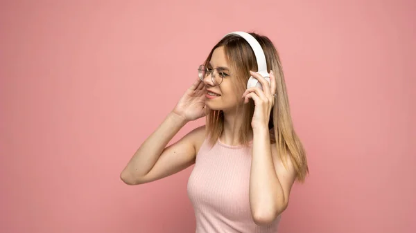 Krásná atraktivní mladá blondýna nosí růžové tričko a brýle v bílých sluchátkách, poslouchá hudbu a usmívá se na růžovém pozadí ve studiu. Uvolňující a užívající si. Životní styl. — Stock fotografie