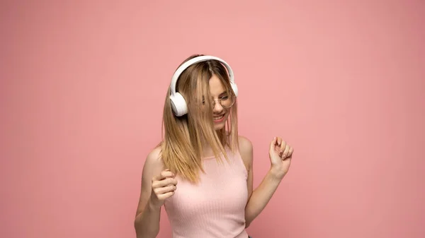 Hermosa mujer rubia joven atractiva con camiseta rosa y gafas en auriculares blancos escuchando música y bailando sobre fondo rosa en el estudio. Relajarse y disfrutar. Estilo de vida. —  Fotos de Stock