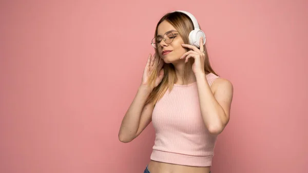 Hermosa mujer rubia joven atractiva con camiseta rosa y gafas en auriculares blancos escuchando música y sonriendo sobre fondo rosa en el estudio. Relajarse y disfrutar. Estilo de vida. —  Fotos de Stock
