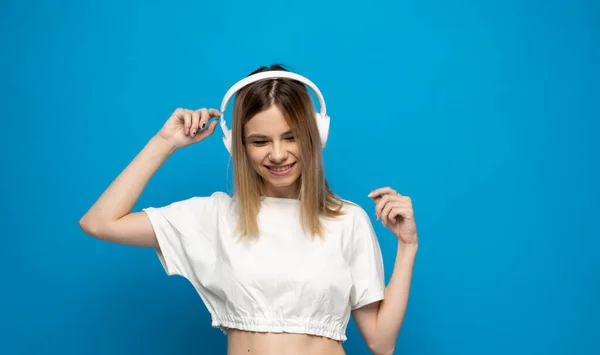 Mooie aantrekkelijke jonge blonde vrouw met een wit t-shirt en een bril in een witte hoofdtelefoon die muziek luistert en danst op een blauwe achtergrond in de studio. Ontspannen en genieten. Levensstijl. — Stockfoto