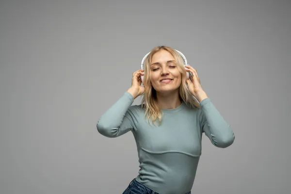 Hermosa mujer rubia joven atractiva con camiseta azul y gafas en auriculares blancos escuchando música y sonriendo sobre fondo gris en el estudio. Relajarse y disfrutar. Estilo de vida. —  Fotos de Stock