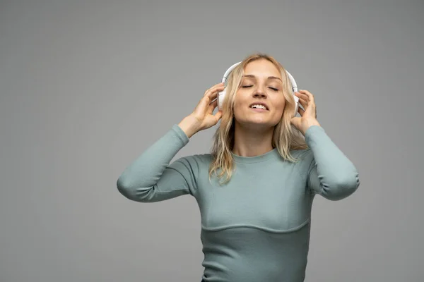 Hermosa mujer rubia joven atractiva con camiseta azul y gafas en auriculares blancos escuchando música y sonriendo sobre fondo gris en el estudio. Relajarse y disfrutar. Estilo de vida. —  Fotos de Stock