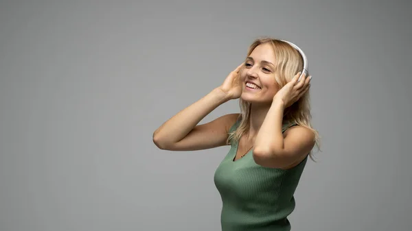 Hermosa mujer rubia joven atractiva con camiseta verde y gafas en auriculares blancos escuchando música y sonriendo sobre fondo gris en el estudio. Relajarse y disfrutar. Estilo de vida. —  Fotos de Stock