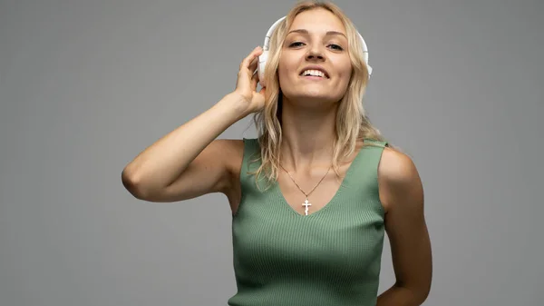 Hermosa mujer rubia joven atractiva con camiseta verde y gafas en auriculares blancos escuchando música y sonriendo sobre fondo gris en el estudio. Relajarse y disfrutar. Estilo de vida. —  Fotos de Stock