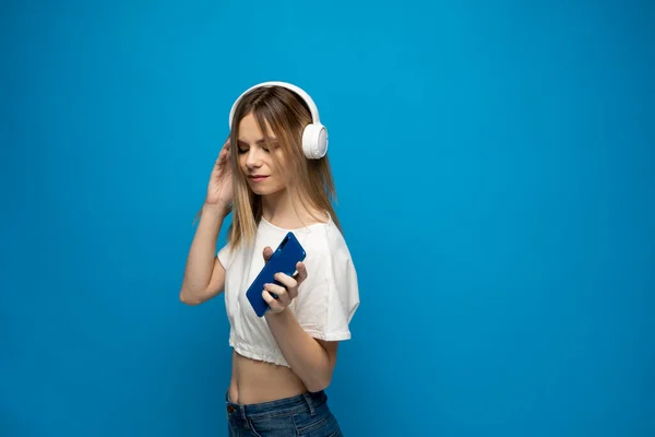 Krásná atraktivní mladá blondýna v bílém tričku a brýlích v bílých sluchátkách, poslouchající hudbu ze smartphonu, tančící a smějící se na modrém pozadí ve studiu. Životní styl. — Stock fotografie