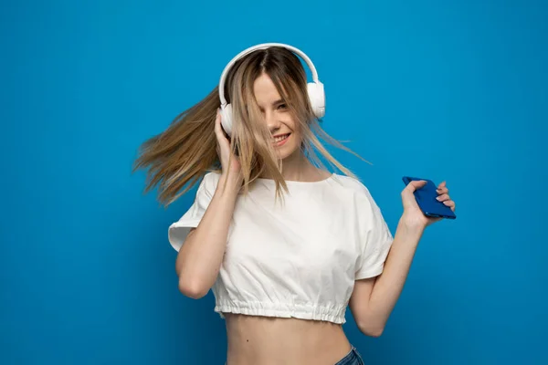 Hermosa mujer rubia joven atractiva con camiseta blanca y gafas en auriculares blancos escuchando música desde el teléfono inteligente, bailando y riendo sobre fondo azul en el estudio. Estilo de vida. —  Fotos de Stock