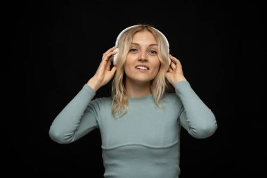 Mavi tişört ve gözlüklü, beyaz kulaklıklı, müzik dinleyen ve stüdyoda siyah arka planda gülümseyen güzel sarışın kadın. Rahatlamak ve eğlenmek. Yaşam biçimi.