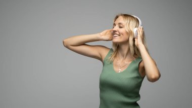 Yeşil tişört ve gözlüklü, beyaz kulaklıklı, müzik dinleyen ve stüdyoda gri arka planda gülümseyen güzel sarışın kadın. Rahatlamak ve eğlenmek. Yaşam biçimi.
