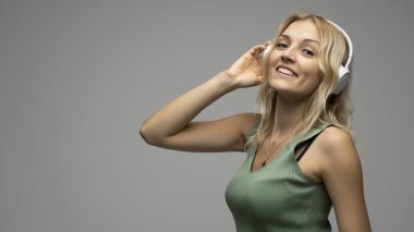 Yeşil tişört ve gözlüklü, beyaz kulaklıklı, müzik dinleyen ve stüdyoda gri arka planda gülümseyen güzel sarışın kadın. Rahatlamak ve eğlenmek. Yaşam biçimi.
