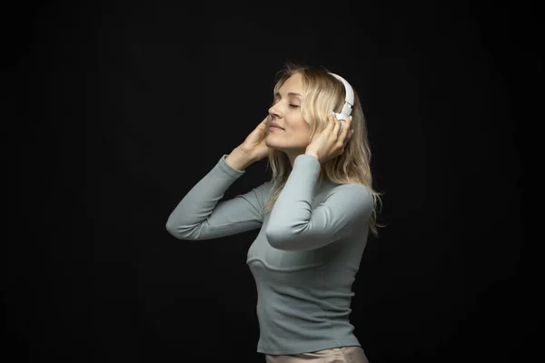 Piękna, atrakcyjna młoda blondynka w niebieskiej koszulce i okularach w białych słuchawkach słuchająca muzyki i uśmiechająca się na czarnym tle w studio. Relaks i przyjemność. Styl życia. — Zdjęcie stockowe