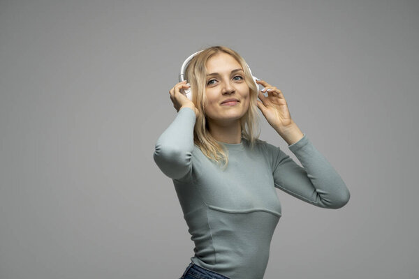 Beautiful attractive young blond woman wearing blue t-shirt and glasses in white headphones listening music and smiling on grey background in studio. Relaxing and enjoying. Lifestyle.