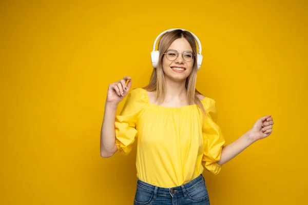 Gyönyörű vonzó, fiatal szőke nő sárga pólóban és szemüvegben fehér fülhallgatóban zenét hallgat, táncol és kéken nevet a stúdióban. Pihentető és élvezet. Életmód. — Stock Fotó
