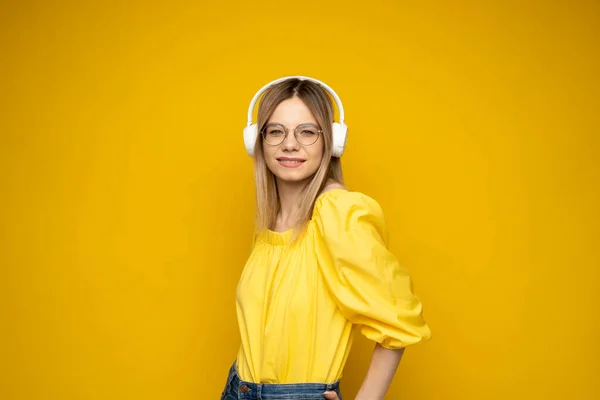Linda jovem mulher loira atraente vestindo camiseta amarela e óculos em fones de ouvido brancos ouvindo música, dançando e rindo em fundo azul no estúdio. Relaxar e desfrutar. Estilo de vida. — Fotografia de Stock