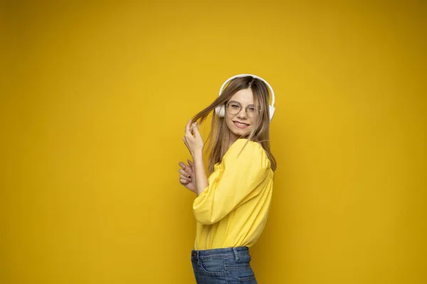 Linda jovem mulher loira atraente vestindo camiseta amarela e óculos em fones de ouvido brancos ouvindo música, dançando e rindo em fundo azul no estúdio. Relaxar e desfrutar. Estilo de vida. — Fotografia de Stock
