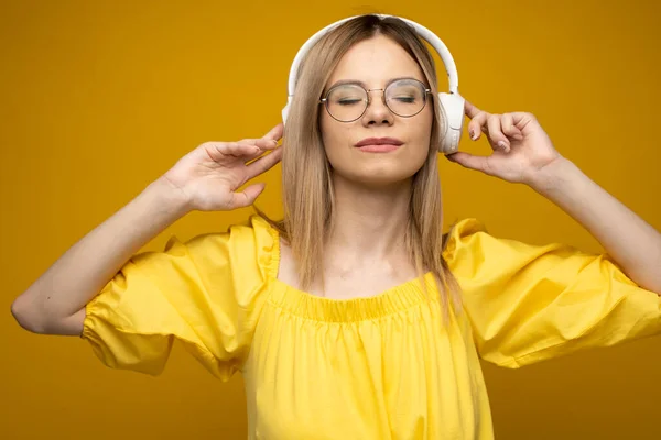 Hermosa mujer rubia joven atractiva con camiseta amarilla y gafas en auriculares blancos escuchando música, bailando y riendo sobre fondo azul en el estudio. Relajarse y disfrutar. Estilo de vida. —  Fotos de Stock