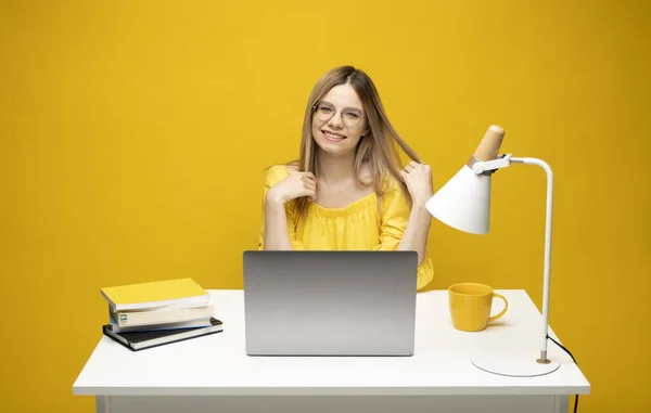 Retrato de una joven sonriente que estudia mientras está sentada en la mesa con una computadora portátil gris, un cuaderno. Mujer de negocios trabajando con un portátil aislado sobre un fondo amarillo. — Foto de Stock