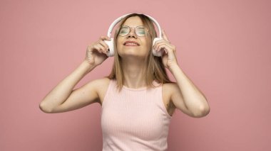 Pembe tişört giyen, beyaz kulaklıklı gözlüklü, müzik dinleyen ve stüdyoda pembe arka planda gülümseyen güzel sarışın bir kadın. Rahatlamak ve eğlenmek. Yaşam biçimi.
