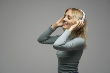 Mavi tişört ve gözlüklü, beyaz kulaklıklı, müzik dinleyen ve stüdyoda gri arka planda gülümseyen güzel sarışın kadın. Rahatlamak ve eğlenmek. Yaşam biçimi.