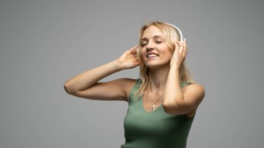 Yeşil tişört ve gözlüklü, beyaz kulaklıklı, müzik dinleyen ve stüdyoda gri arka planda gülümseyen güzel sarışın kadın. Rahatlamak ve eğlenmek. Yaşam biçimi.