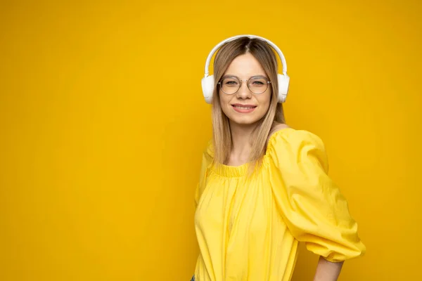 Krásná atraktivní mladá blondýnka ve žlutém tričku a brýlích v bílých sluchátkách, poslouchající hudbu, tančící a smějící se na modrém pozadí ve studiu. Uvolňující a užívající si. Životní styl. — Stock fotografie
