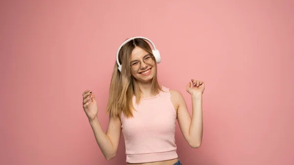 Linda jovem mulher loira atraente vestindo camiseta rosa e óculos em fones de ouvido brancos ouvindo música e dançando em fundo rosa em estúdio. Relaxar e desfrutar. Estilo de vida. — Fotografia de Stock