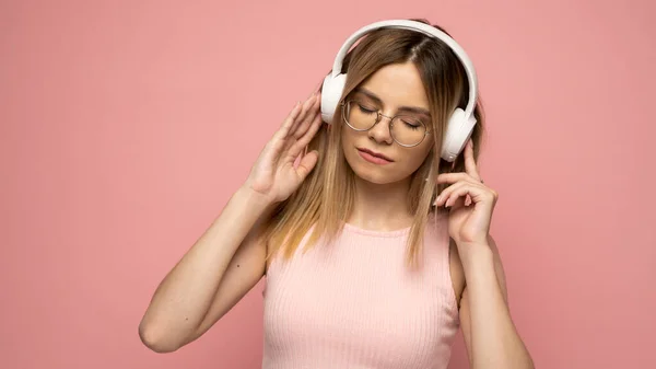 Krásná atraktivní mladá blondýna nosí růžové tričko a brýle v bílých sluchátkách, poslouchá hudbu a usmívá se na růžovém pozadí ve studiu. Uvolňující a užívající si. Životní styl. — Stock fotografie
