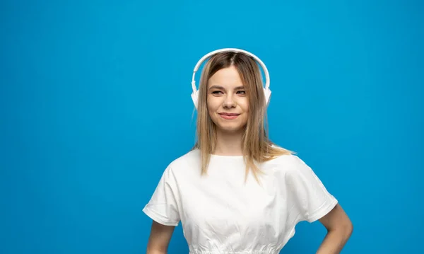 Linda jovem mulher loira atraente vestindo camiseta branca e óculos em fones de ouvido brancos ouvindo música e sorrindo em fundo azul no estúdio. Relaxar e desfrutar. Estilo de vida. — Fotografia de Stock