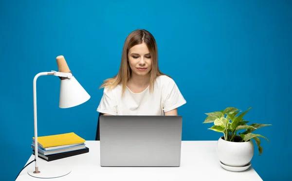 Retrato de uma jovem bonita estudando enquanto se senta na mesa com computador portátil cinza, notebook. Sorrindo mulher de negócios trabalhando com um laptop isolado em um fundo azul. — Fotografia de Stock