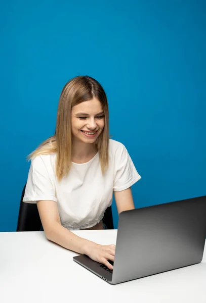 Portrait of a pretty young woman studying while sitting at the table with grey laptop computer, notebook. Smiling business woman working with a laptop isolated on a blue background. — Stock Photo, Image