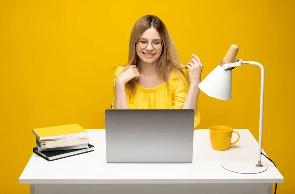 Retrato de una joven sonriente que estudia mientras está sentada en la mesa con una computadora portátil gris, un cuaderno. Mujer de negocios trabajando con un portátil aislado sobre un fondo amarillo. — Foto de Stock