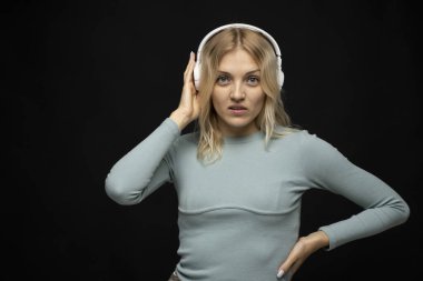 Mavi tişört ve gözlüklü, beyaz kulaklıklı, müzik dinleyen ve stüdyoda siyah arka planda gülümseyen güzel sarışın kadın. Rahatlamak ve eğlenmek. Yaşam biçimi.