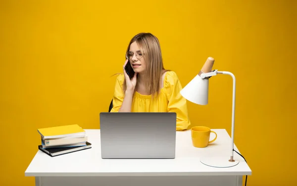Młoda atrakcyjna biznesmenka z blond włosami siedząca przy stole, pracująca nad laptopem i rozmawiająca przez telefon z klientem. Młoda, wesoła uczennica rozmawiająca ze smartfonem. Studiowanie. — Zdjęcie stockowe