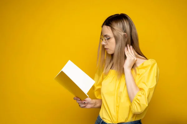 Умная молодая девушка держит в руках книгу и читает ее на желтом фоне. Портрет привлекательной женщины в жёлтой блузке и в очках, читающей книгу. Образование, учеба, знания. — стоковое фото