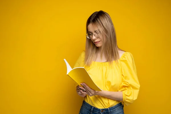 Умная молодая девушка держит в руках книгу и читает ее на желтом фоне. Портрет привлекательной женщины в жёлтой блузке и в очках, читающей книгу. Образование, учеба, знания. — стоковое фото